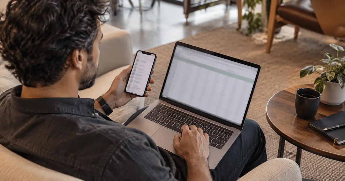 Pessoa compara uma planilha no notebook com um aplicativo no celular em uma área de trabalho.